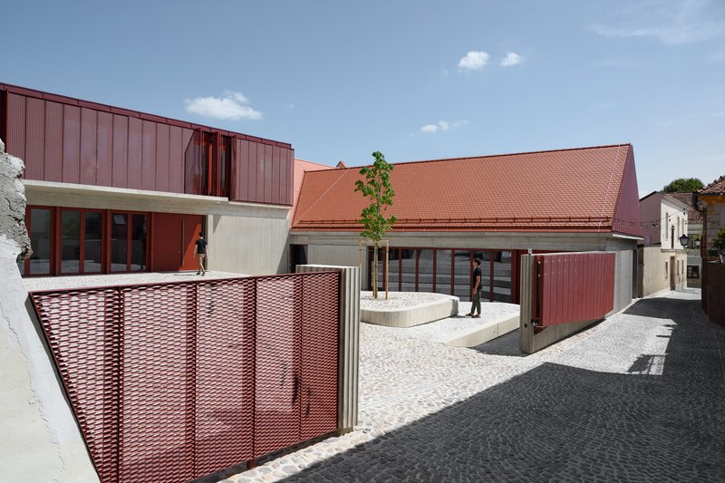 Cobblestone courtyard showing perforated red metal screen fence and sloped tile roof under blue sky