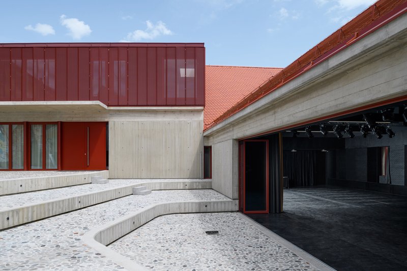 Courtyard with terrazzo paving and sunken concrete seating steps facing red metal cladding and orange tile roof