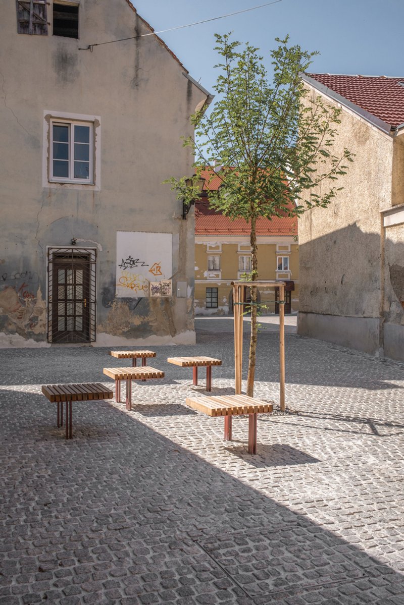 Cobblestone plaza with timber and metal seating elements around a young tree and weathered stucco facades