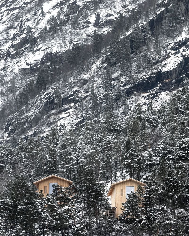 Two timber cabins nestled among pine trees against a steep rocky mountainside dusted with snow