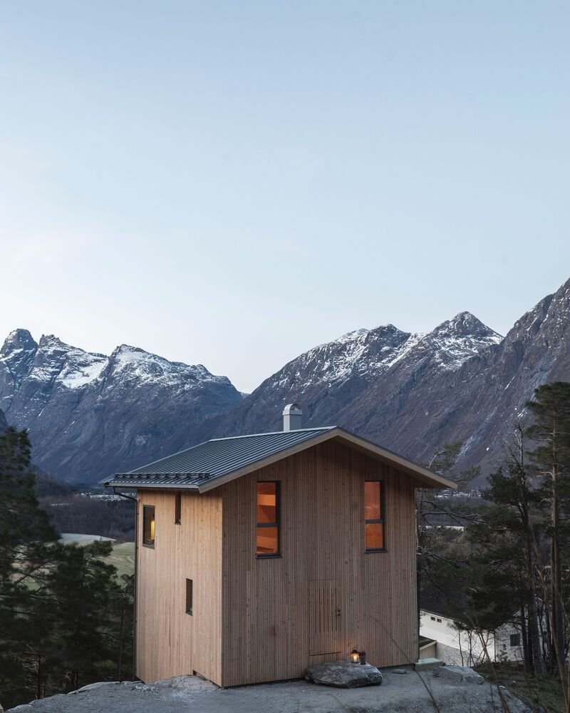Vertical timber facade with illuminated windows at twilight framed by evergreen trees and distant peaks