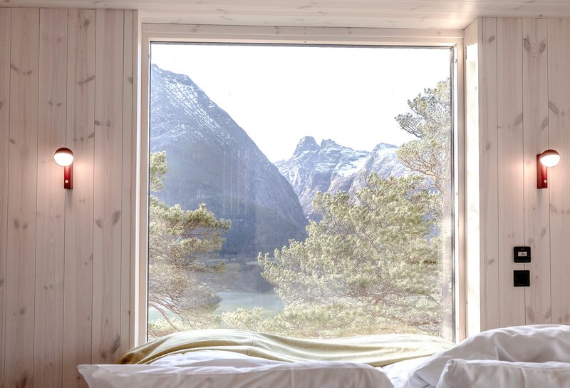Bedroom window framing snowcapped mountains and riverside trees through light wood paneling with wall sconces