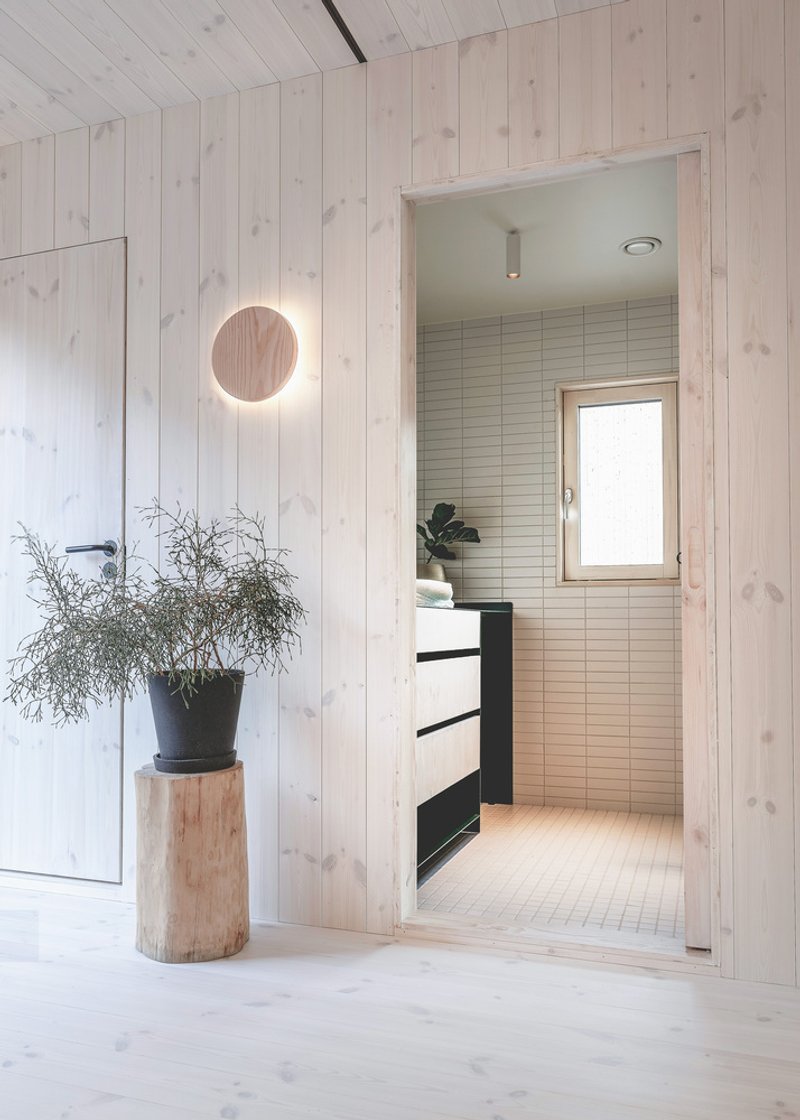 Interior hallway with circular wall light and potted plant leading to bathroom with horizontal tile