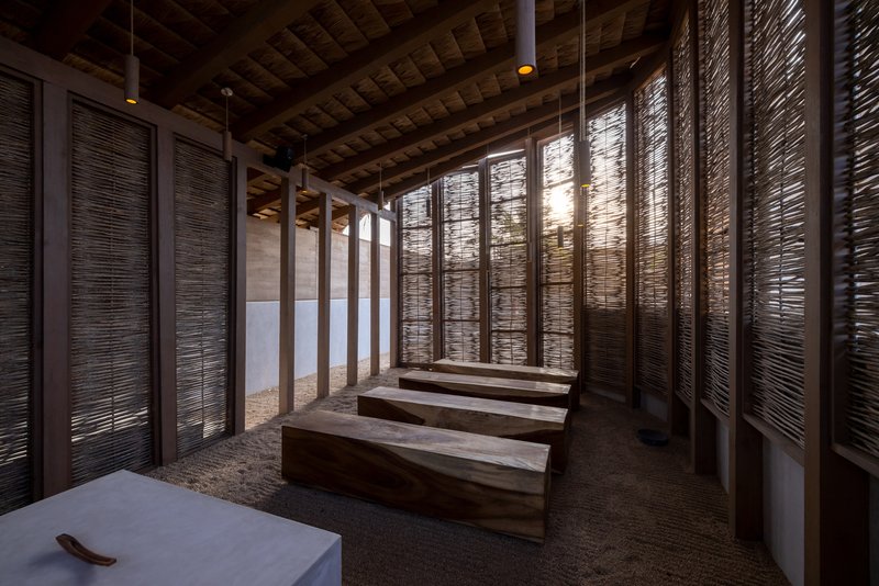 Timber benches arranged in rows beneath reed ceiling and backlit woven screen panels at dusk