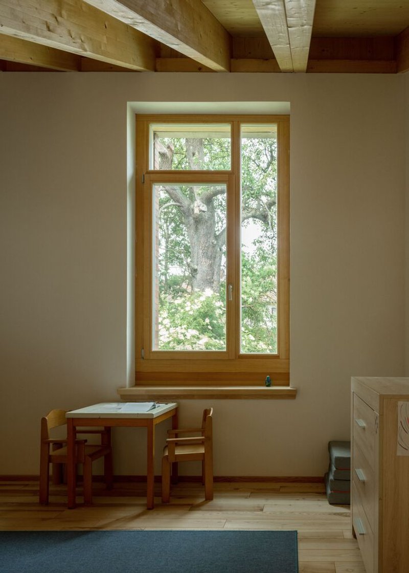 Tall timber-framed window with deep reveal overlooking trees, paired with a children's table