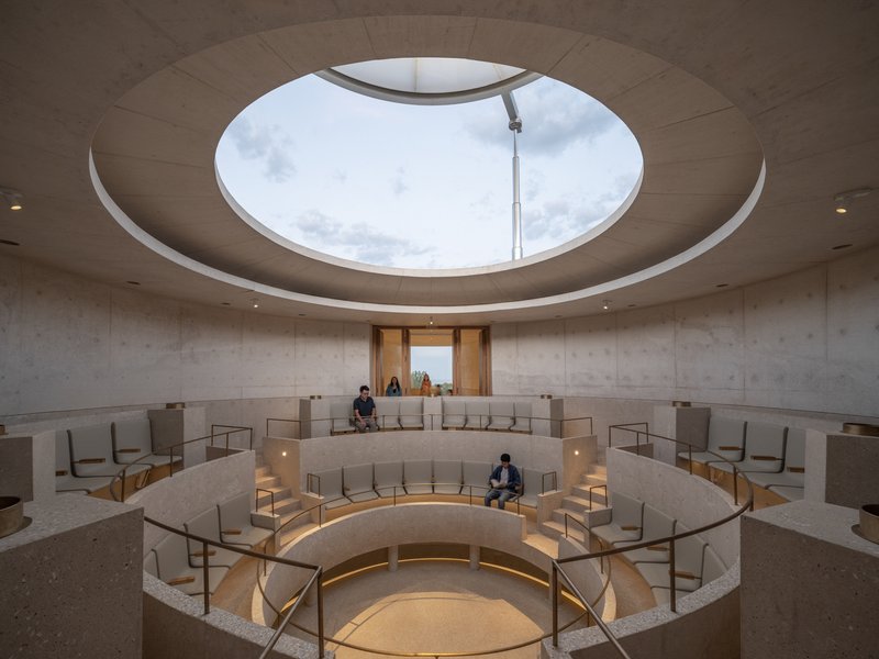 Interior circular sanctuary with tiered seating and central oculus opening to cloudy sky