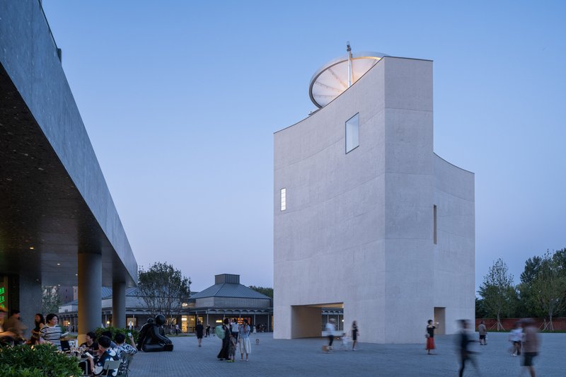 White concrete tower with vertical slit windows and rooftop disc in blue evening light