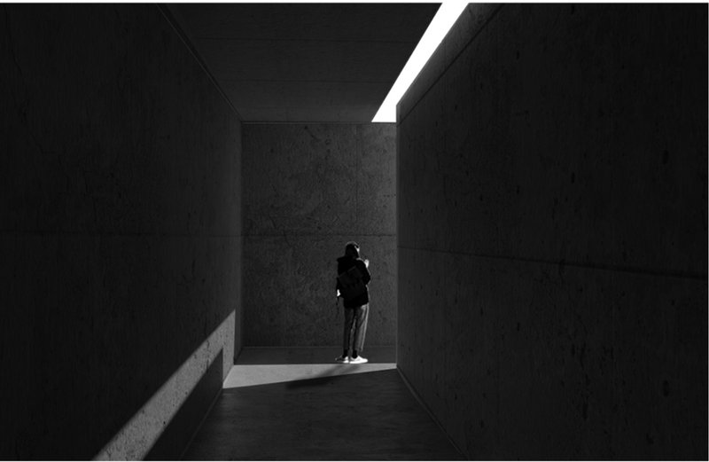 Visitor standing in a concrete chamber beneath a slender skylight casting angular shadows