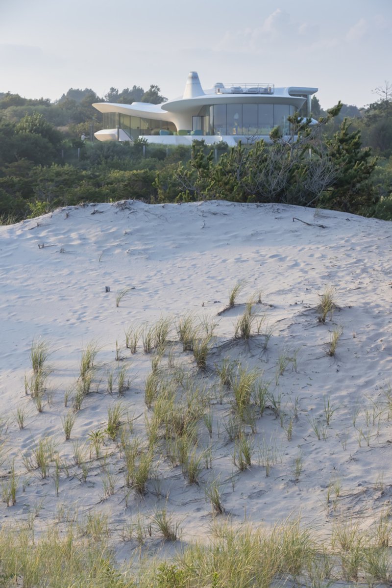 White curved volume with glass walls and cantilevered terraces rising above a sand dune with scattered beach grasses