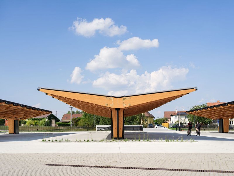 Timber canopy structure with upswept wing forms over a paved plaza with cyclists passing