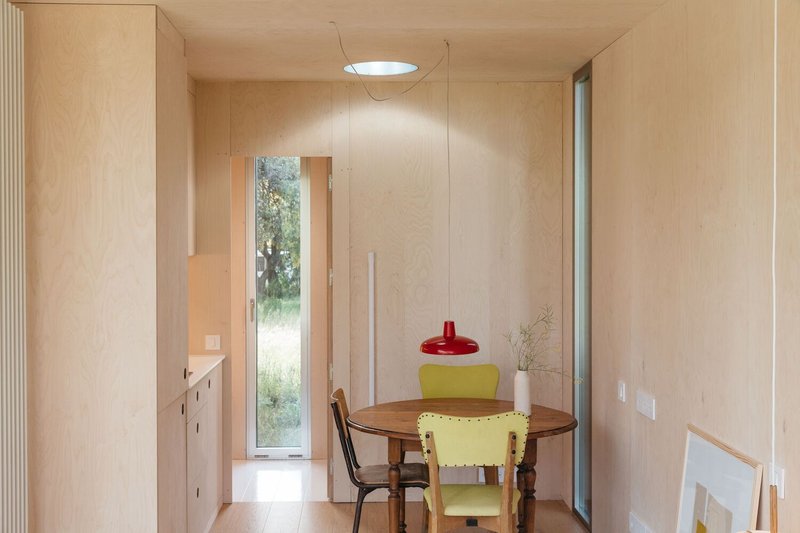 Light plywood-lined dining alcove with round table and narrow window opening to landscape beyond