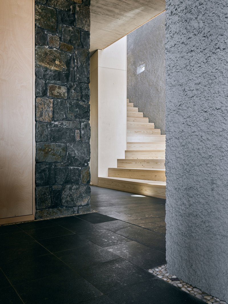 Light wood staircase rising between dark stone wall and pebble-lined textured concrete surface