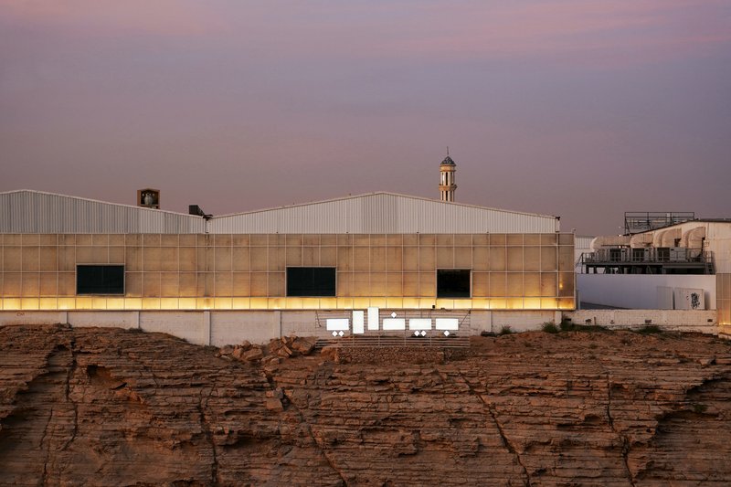 Illuminated translucent facade with rectangular window openings above layered sandstone cliffs at dusk