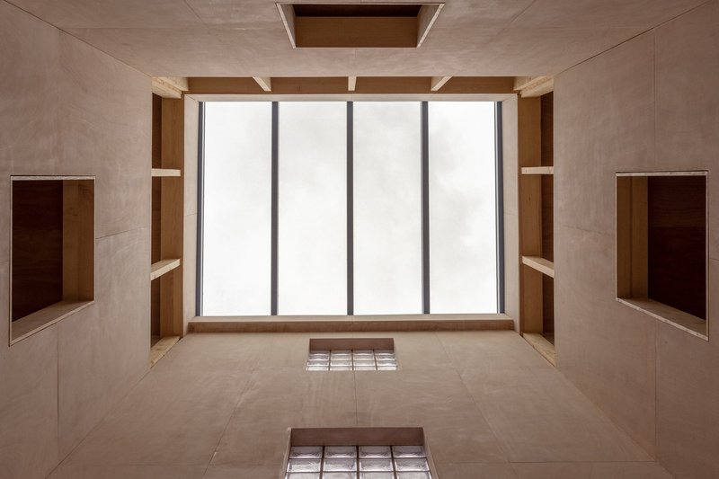 Looking up at a plywood-clad shaft with tall glazing and recessed timber niches on both sides
