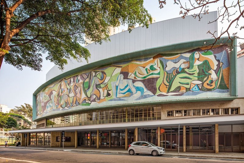 Street facade with curved canopy bearing colorful figural mosaic beneath overhanging trees at golden hour