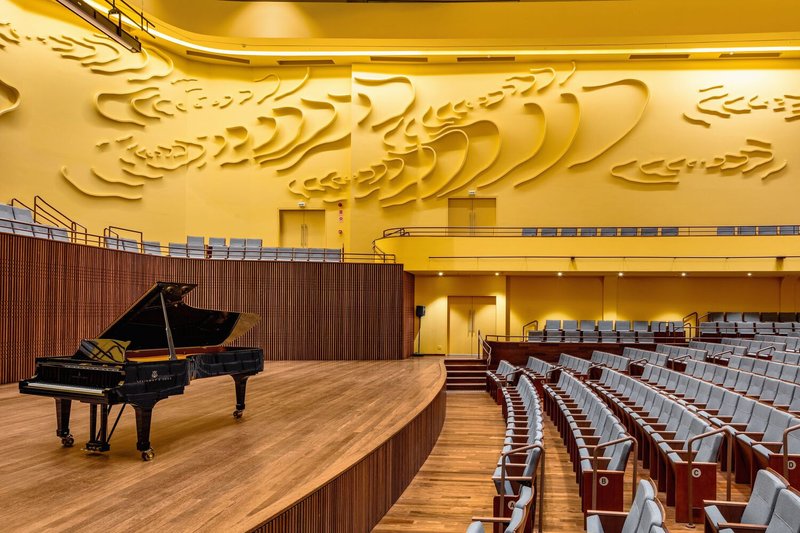 Grand piano on curved timber stage beneath the sculpted yellow wall and auditorium seating under warm lighting