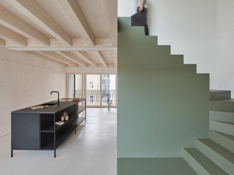 Open kitchen with black island beneath exposed timber joists and a person ascending the sage stair