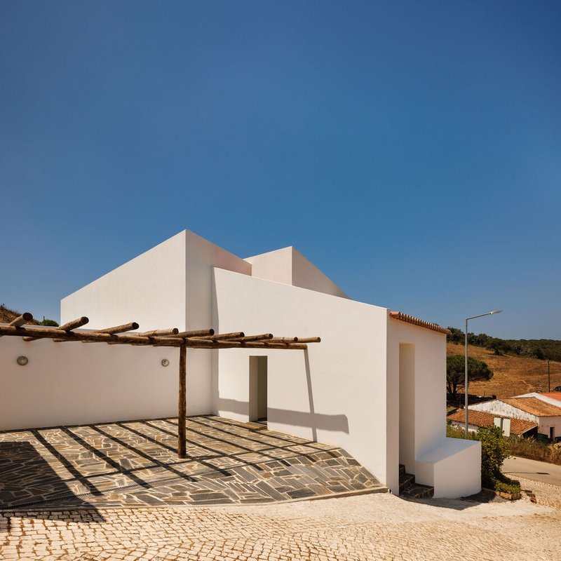 White stucco facade with timber pergola casting shadows over cobbled paving under clear blue sky