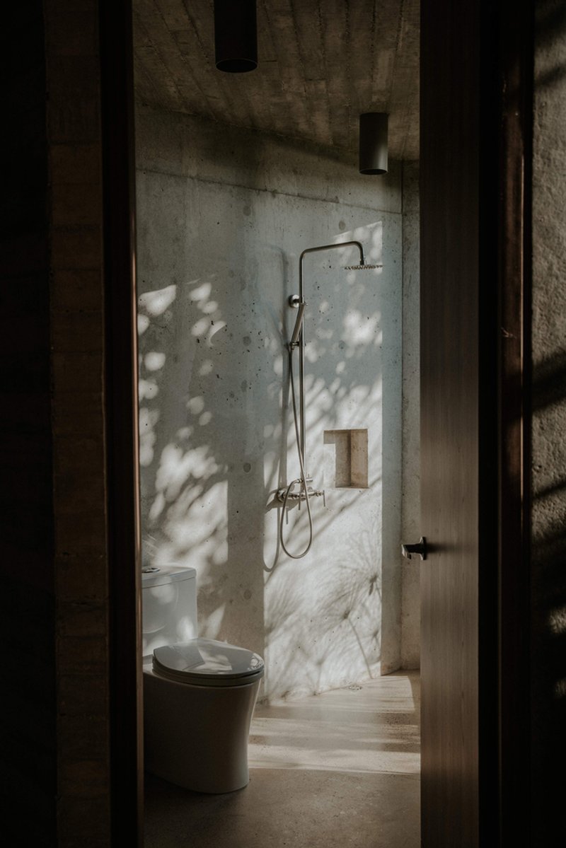 Bathroom doorway framing concrete and marble shower enclosure with dappled sunlight filtering through adjacent foliage