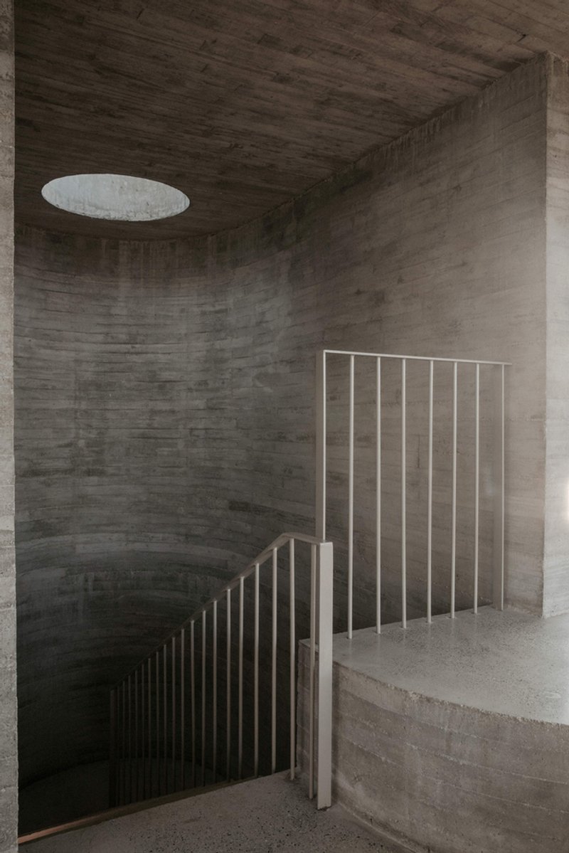 Curved concrete stairwell with vertical metal balustrade and circular skylight above board-formed walls