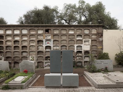 This image depicts a cemetery with a large brick mausoleum wall featuring numerous arched niches. In