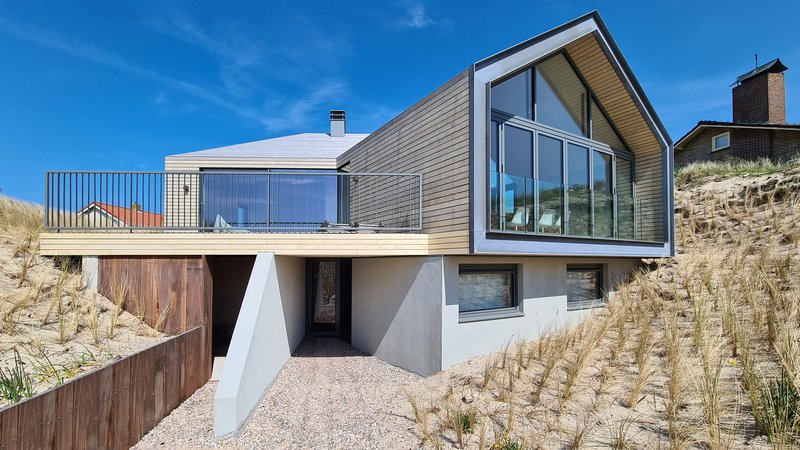 Angled facade with horizontal timber cladding and glazed gable end set into sandy hillside