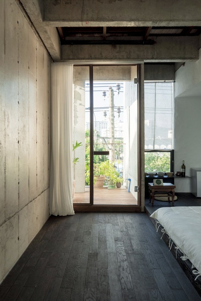 Bedroom with board-formed concrete walls, dark timber flooring, and glazed doors opening to a planted balcony