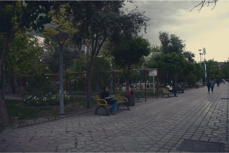 Paved plaza with red metal playground equipment and yellow benches beneath mature trees
