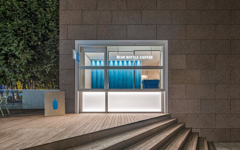 Glass-fronted kiosk with blue corrugated metal wall and timber deck steps at dusk