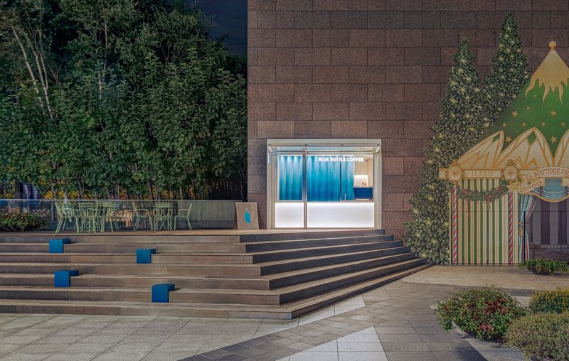 Timber stepped plaza with blue accent cubes leading to a glazed kiosk at night