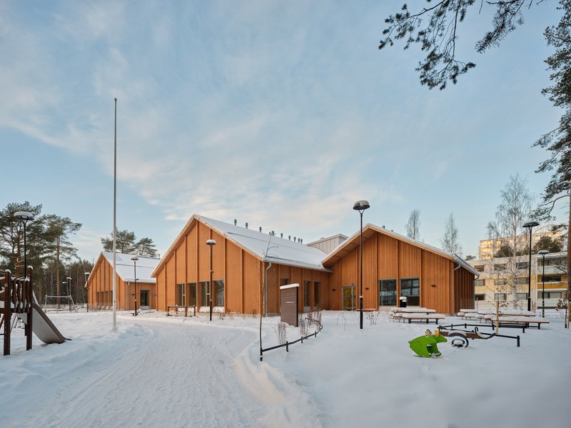 Cluster of timber-clad gabled volumes with playground equipment in a snow-covered clearing at twilight