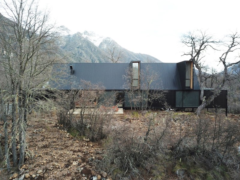 Linear metal-clad volumes on a rocky hillside with bare winter trees and snow-capped mountains beyond