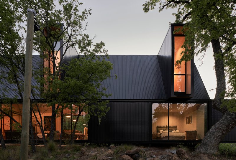 Dark metal-clad volumes with floor-to-ceiling glazing nestled among mature trees at twilight