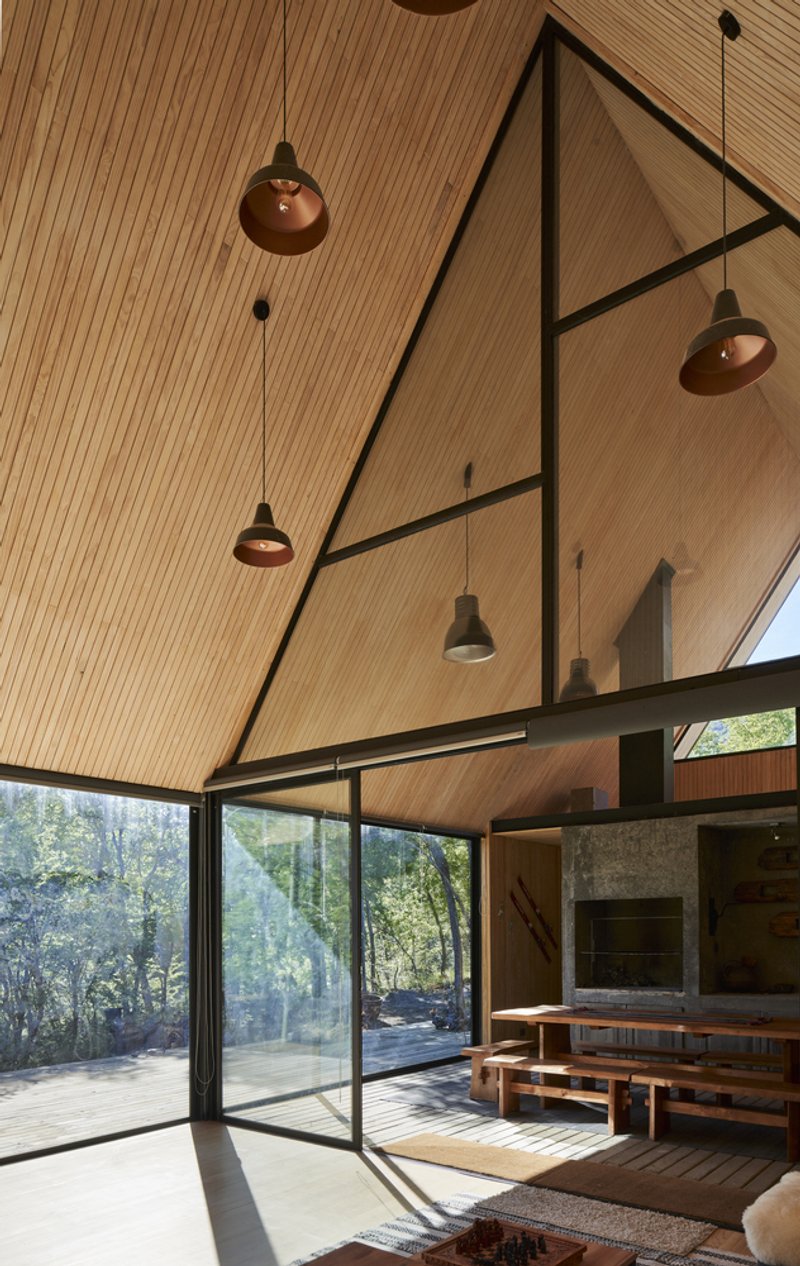 Double-height living space with sloped timber ceiling, pendant lights, and full-height glazing opening to the forest