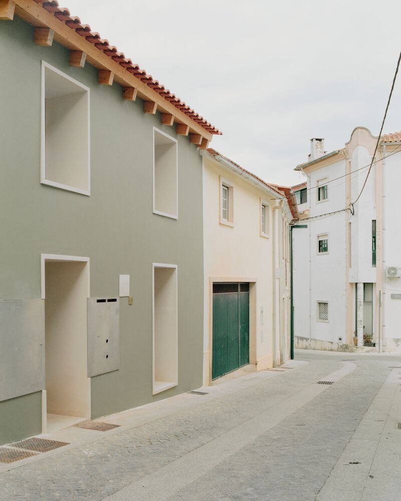 Street facade with deep recessed openings in sage-green render and red clay tile roof