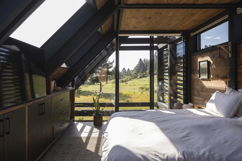 Bedroom with sloped timber ceiling, black steel frame glazing, and morning sunlight across white bedding