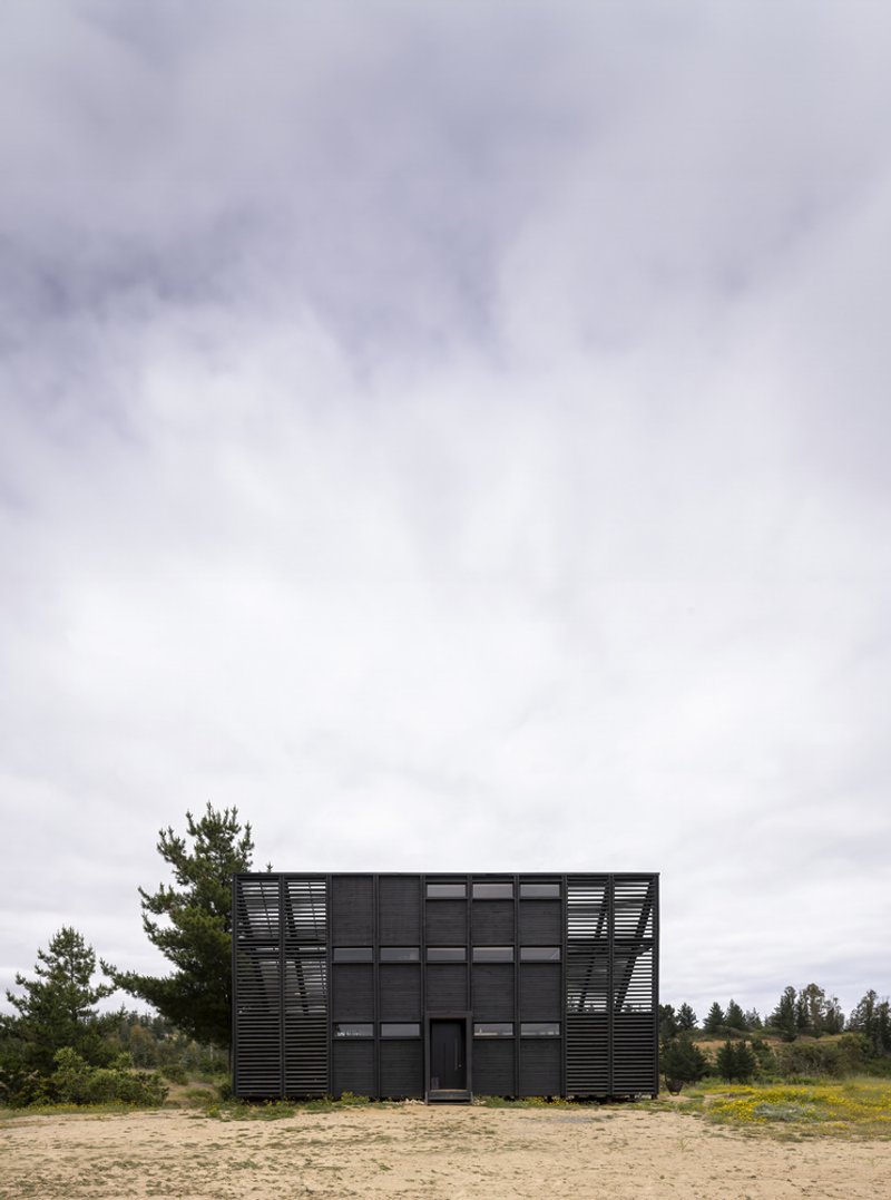 Black corrugated metal facade with horizontal louvers and windows on sandy ground under overcast sky