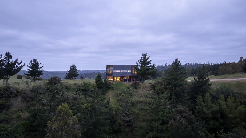 The illuminated interior visible through glazed openings at dusk above forested slope