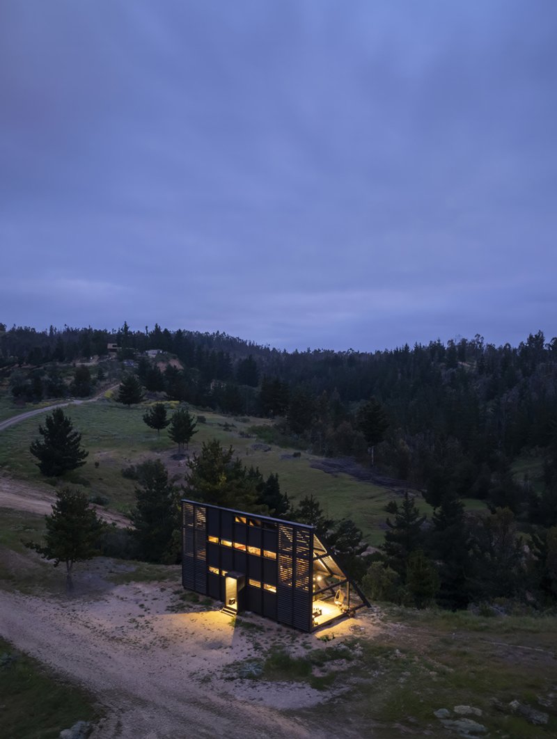 Aerial view of illuminated A-frame cabin on hillside meadow surrounded by forest at twilight