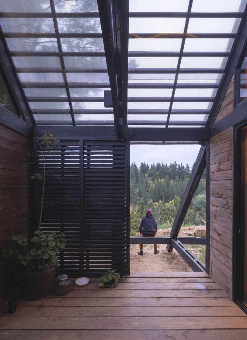 Covered timber deck with translucent roof panels and seated figure overlooking forested hillside