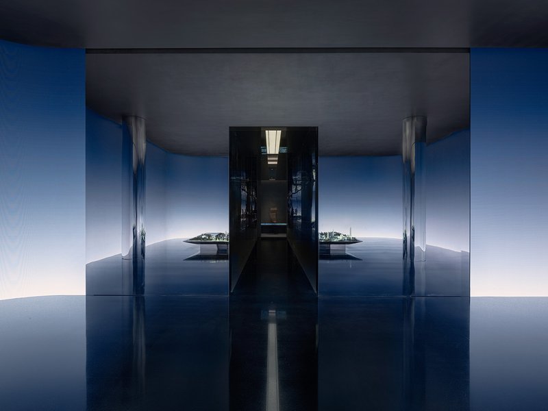 Symmetrical corridor with reflective blue floor and dark ceiling opening to a bright courtyard beyond