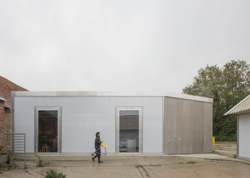 Street facade with white panels and corrugated metal siding as a figure walks past with a yellow crate