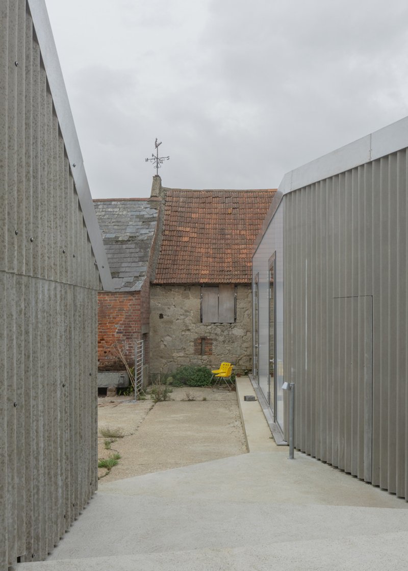 Concrete passage between corrugated metal volumes leading to weathered brick outbuilding with terracotta roof and yellow chair