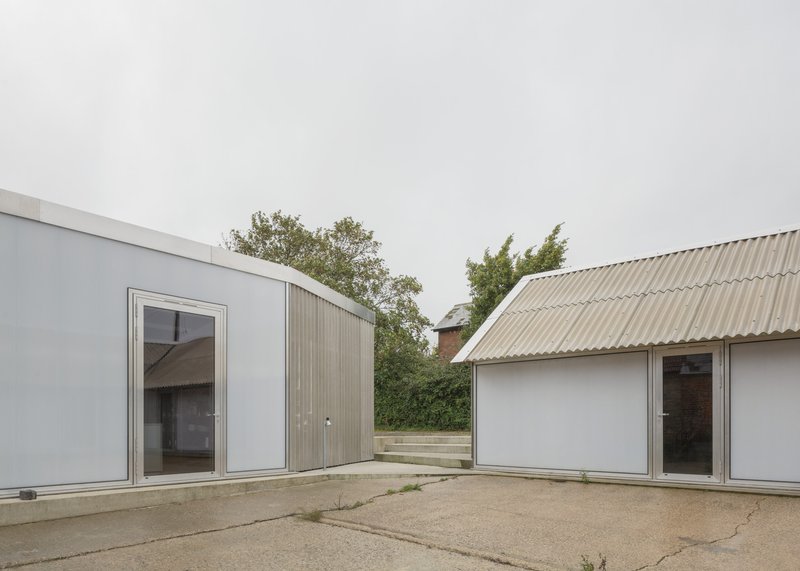 View across concrete courtyard showing corrugated metal roofs and timber cladding with trees visible beyond