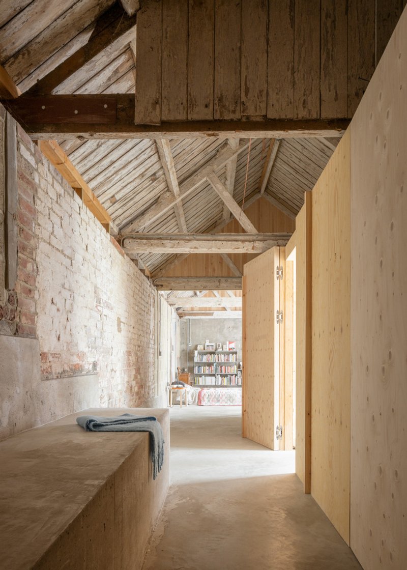 Passage with exposed brick wall and weathered timber roof structure framing view to library shelving beyond