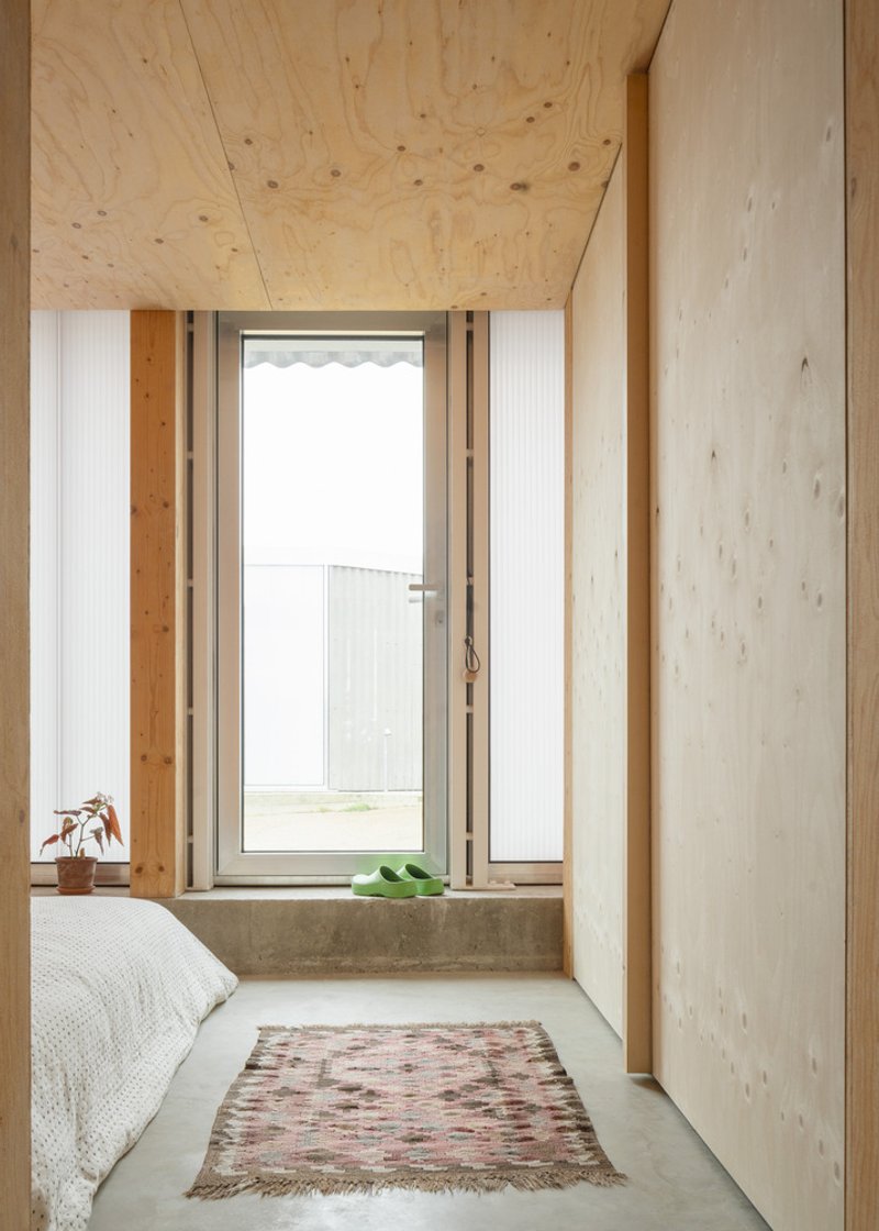 Bedroom corner with glazed door, plywood walls and woven rug on polished concrete floor