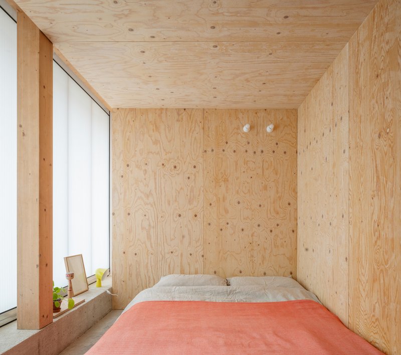 Plywood-lined sleeping alcove with pink duvet and concrete windowsill holding a plant