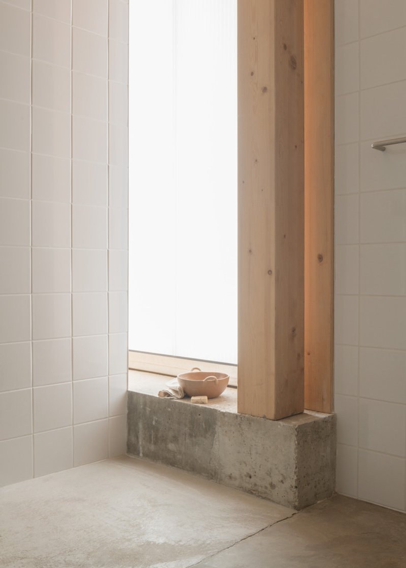 White tiled bathroom corner with tall window and concrete sill holding a wooden bowl