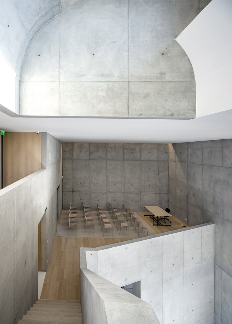 Curving concrete auditorium with board-formed walls and rows of timber seating below vaulted ceiling
