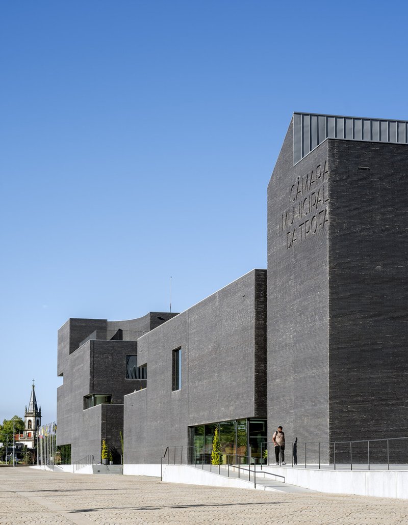Interlocking brick-clad volumes with a person walking near the entrance plaza beside a church spire